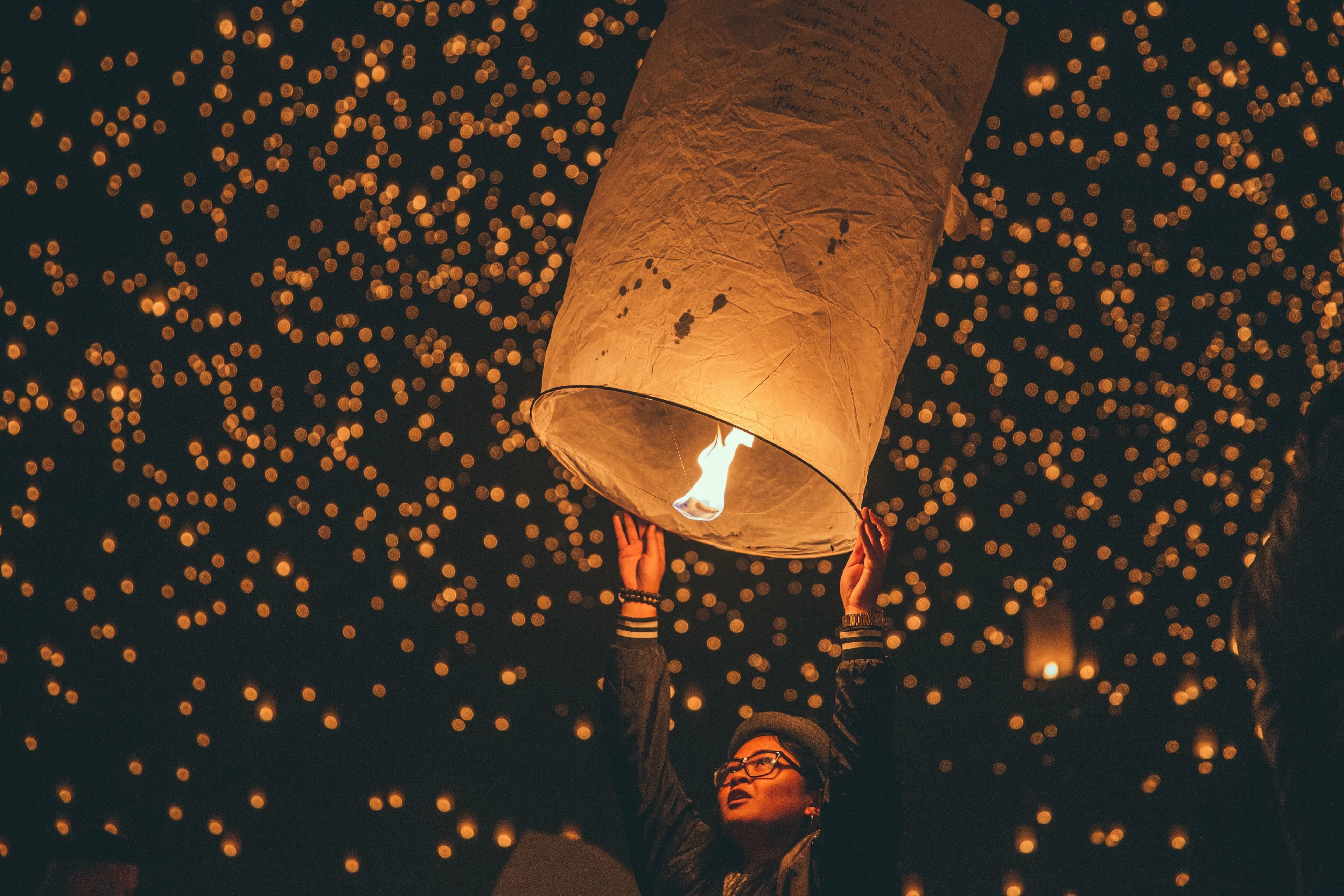 Una persona sostiene un farolillo de papel con luz en su interior, rodeada de numerosas luces pequeñas al fondo.