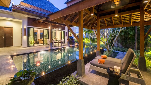 A poolside area with a lounge chair, a table with a candle, and a view of a house with a covered patio. - The Bell Pool Villa Resort (Kamala)