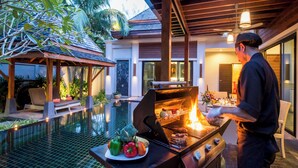 A man grilling outdoors near a pool with a patio area and tropical plants. - The Bell Pool Villa Resort (Kamala)