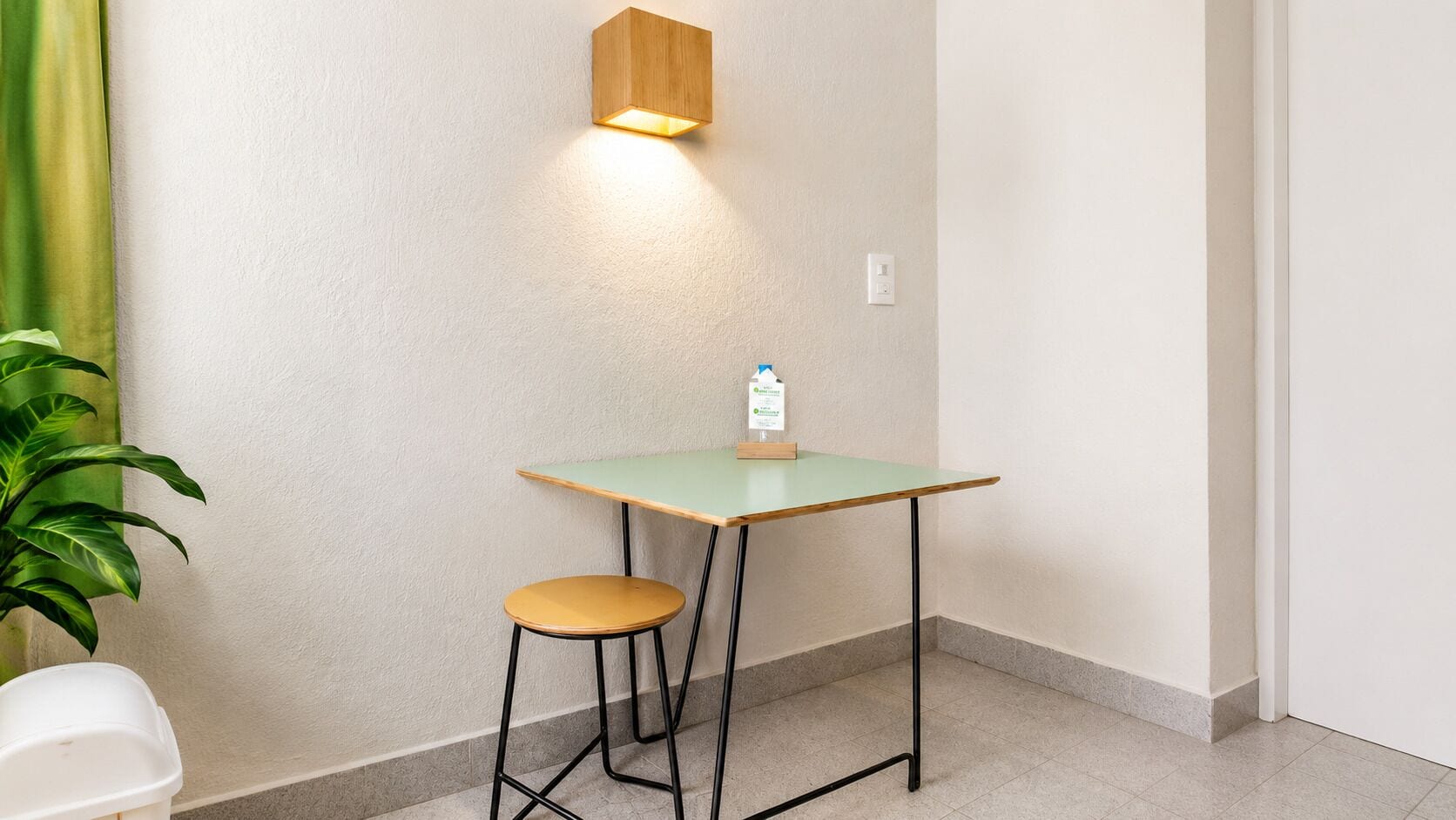 A small room with a table, a stool, a potted plant, and a wall-mounted light fixture.