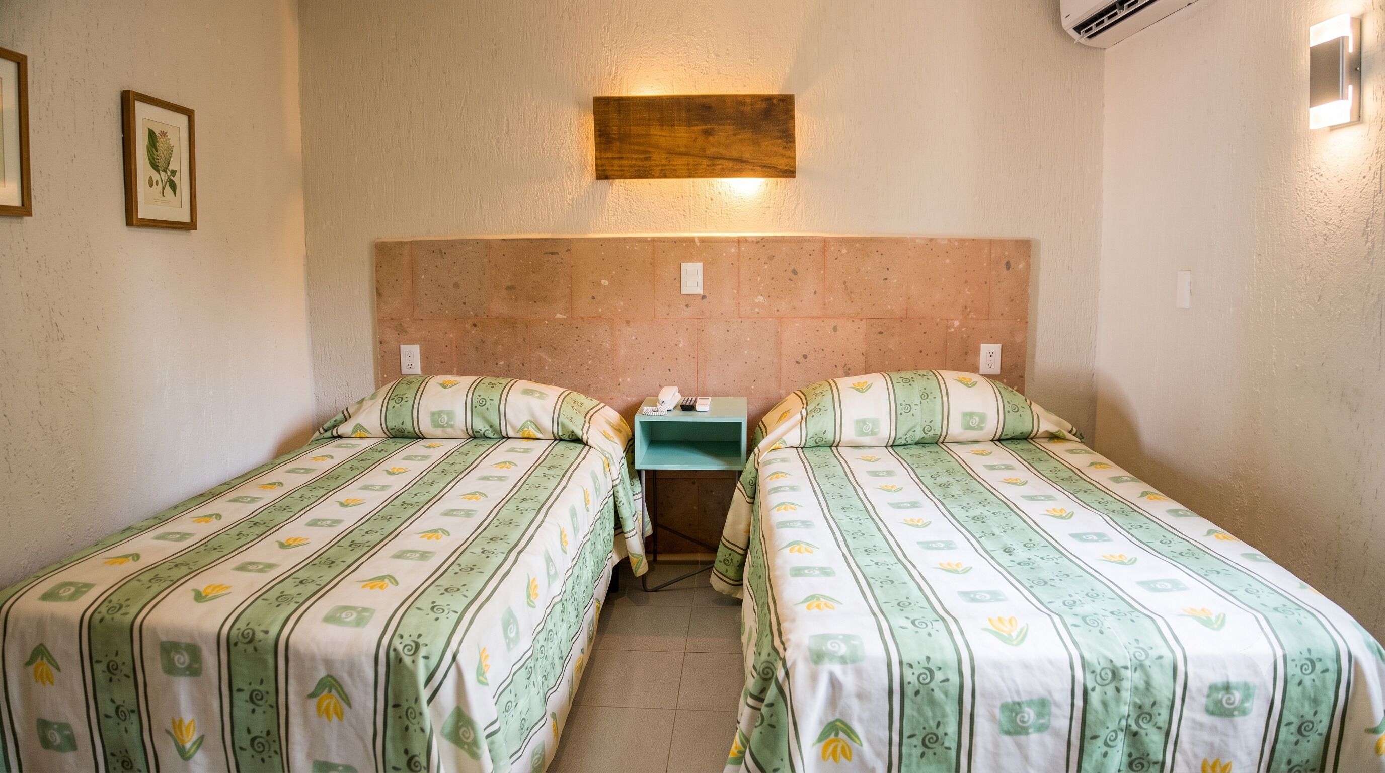 Une chambre avec deux lits, chacun agrémenté de draps à motifs, une petite table de chevet avec un téléphone et un bloc-notes, et un luminaire fixé au mur.