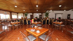 A spacious dining area with wooden tables and chairs, large windows, and potted plants. - Dorados Conventions & Resort (Yautepec)