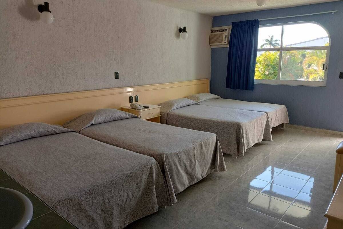 A hotel room with two beds, a small bedside table with a phone, and a window with a view of palm trees.