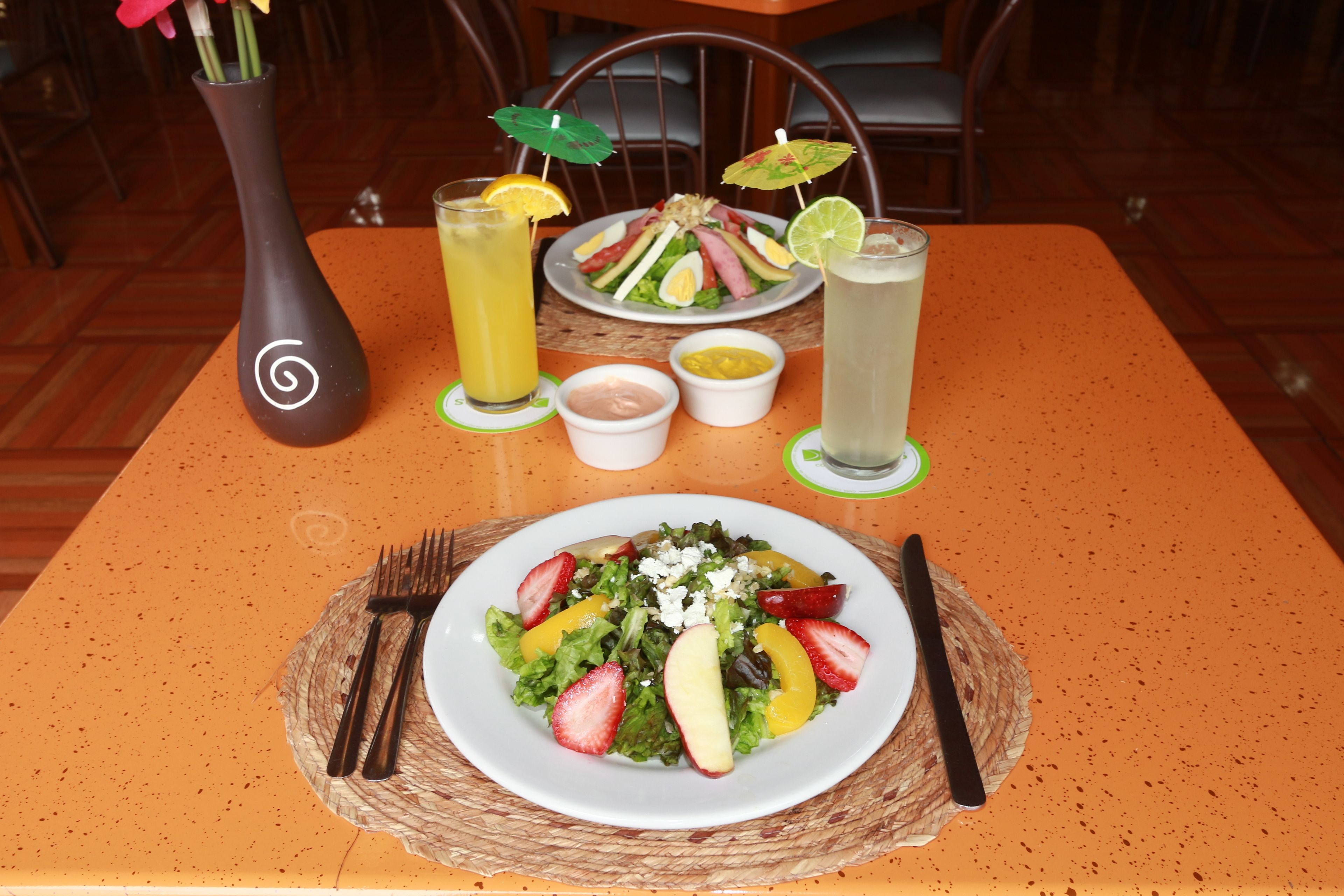 Une table avec une assiette de salade, deux boissons, un vase et des condiments.