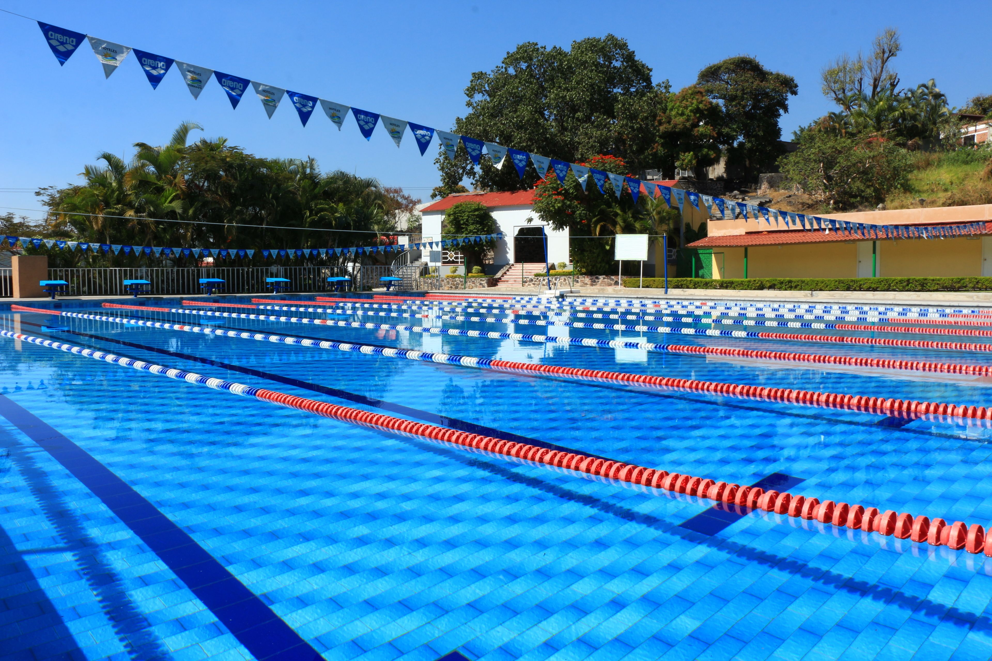 Une piscine dotée de plusieurs couloirs, d’une zone couverte pour les spectateurs et, au fond, un bâtiment avec un toit rouge.