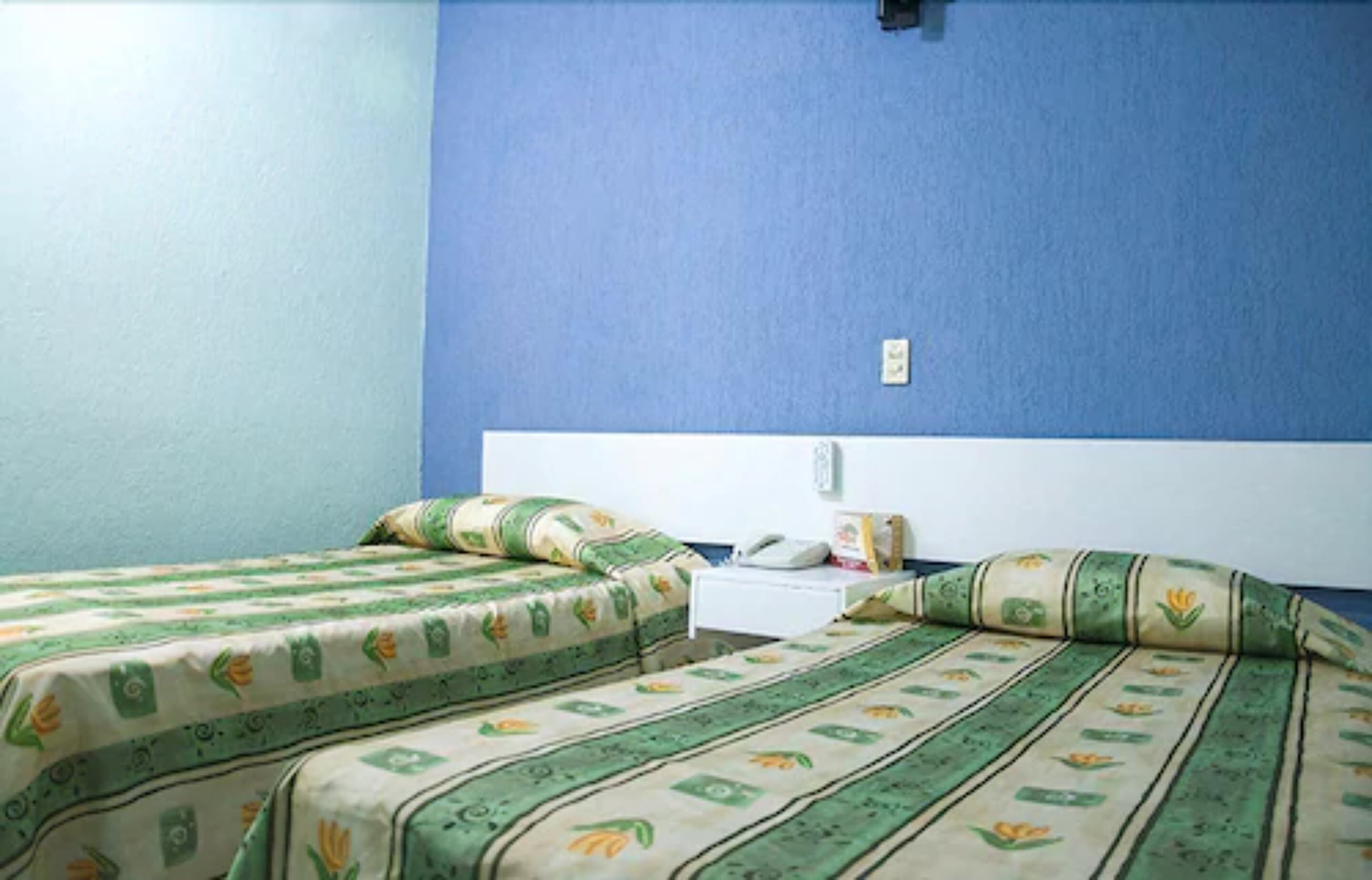 Una habitación con dos camas, cada una con ropa de cama verde y beige, una mesita de noche blanca y una pared azul.