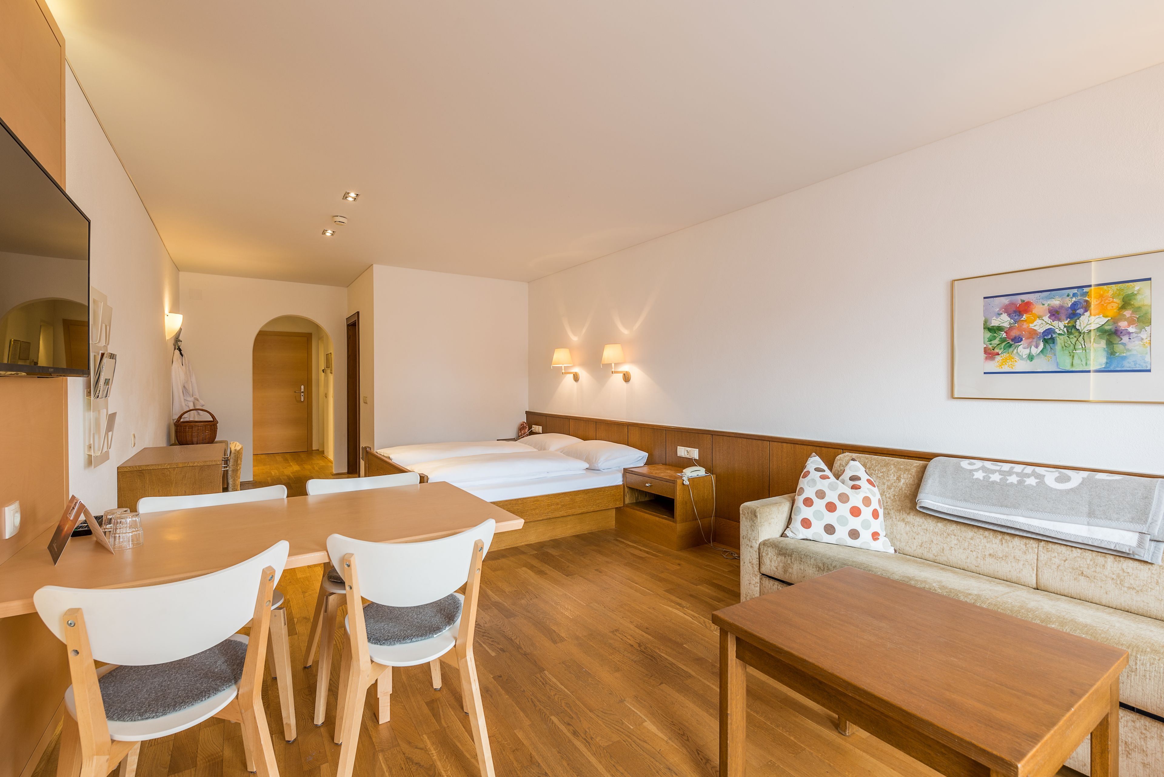 A hotel room with a wooden dining table, two chairs, a sofa, a bed, and a painting on the wall.
