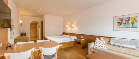 A hotel room with a wooden dining table, two chairs, a sofa, a bed, and a painting on the wall.