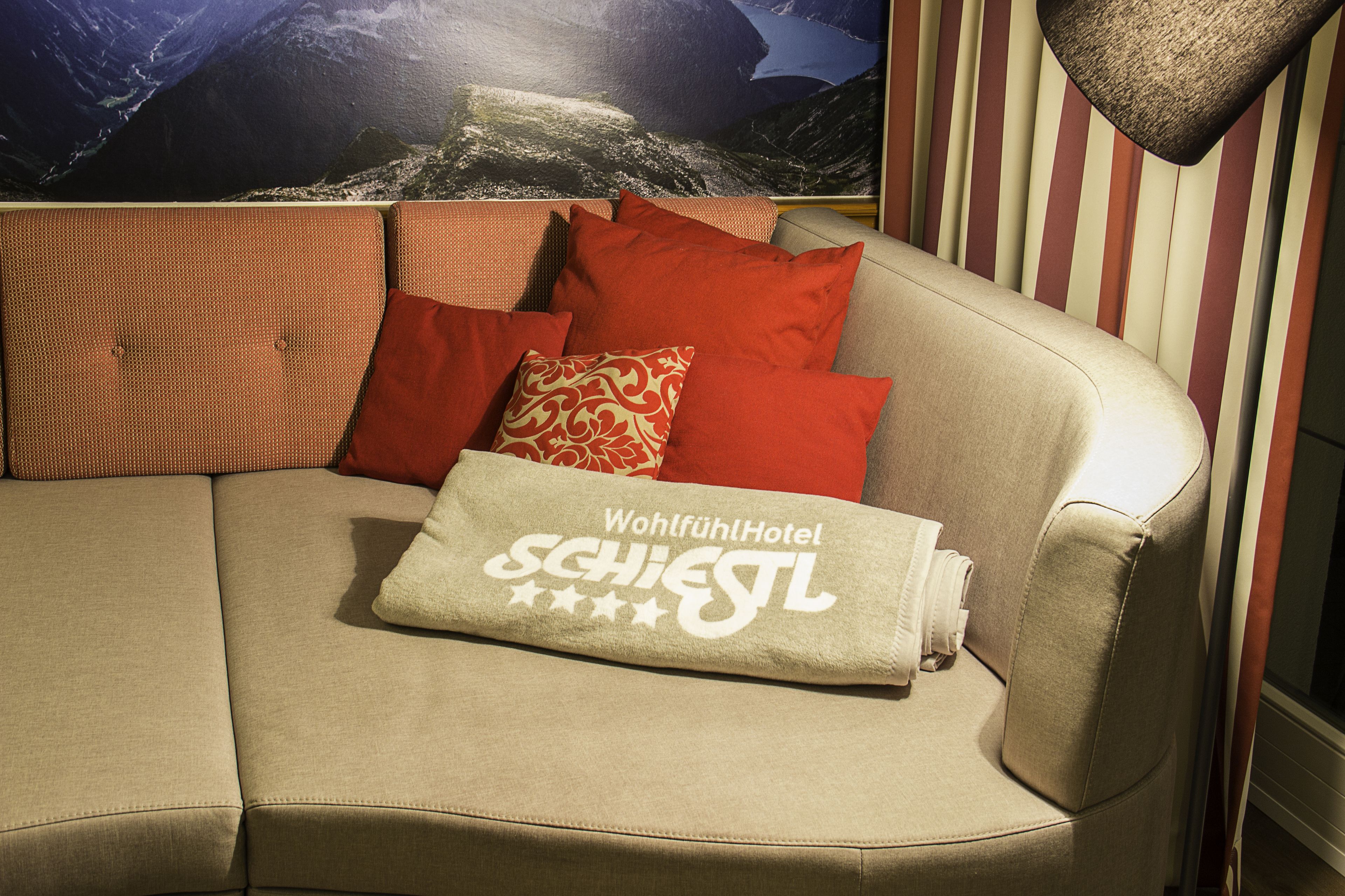 Un sofá color beige con almohadones rojos y estampados, una manta con el texto "Wohlfuht Hotel SCHIEGL", y un mural de pared con un paisaje.
