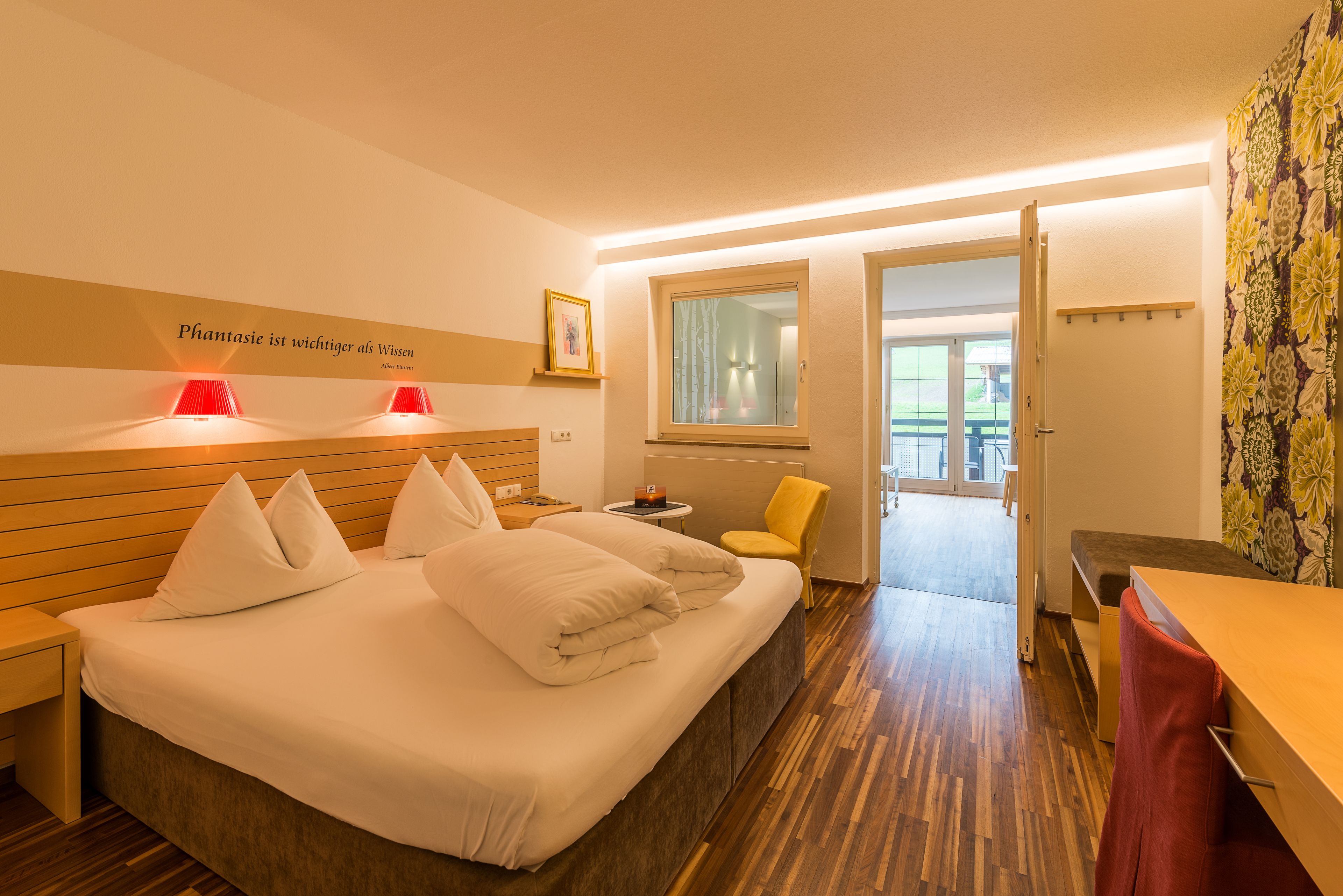 Quarto de hotel com cama, mesas de cabeceira, uma cadeira, escrivaninha e uma janela.