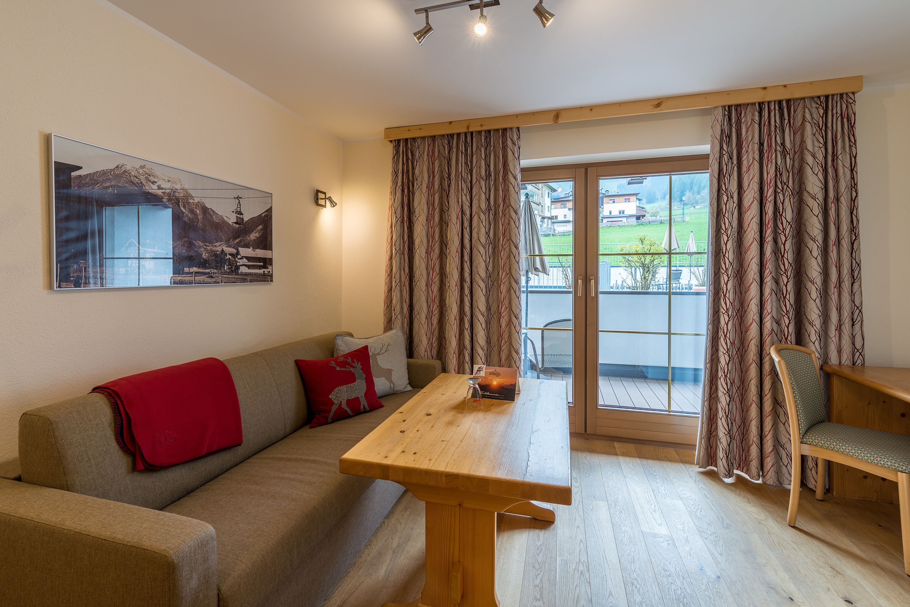 Une pièce avec une table en bois, un canapé recouvert d’une couverture rouge et une chaise. Il y a une fenêtre avec des rideaux, d’où l’on peut voir des bâtiments à l’extérieur.