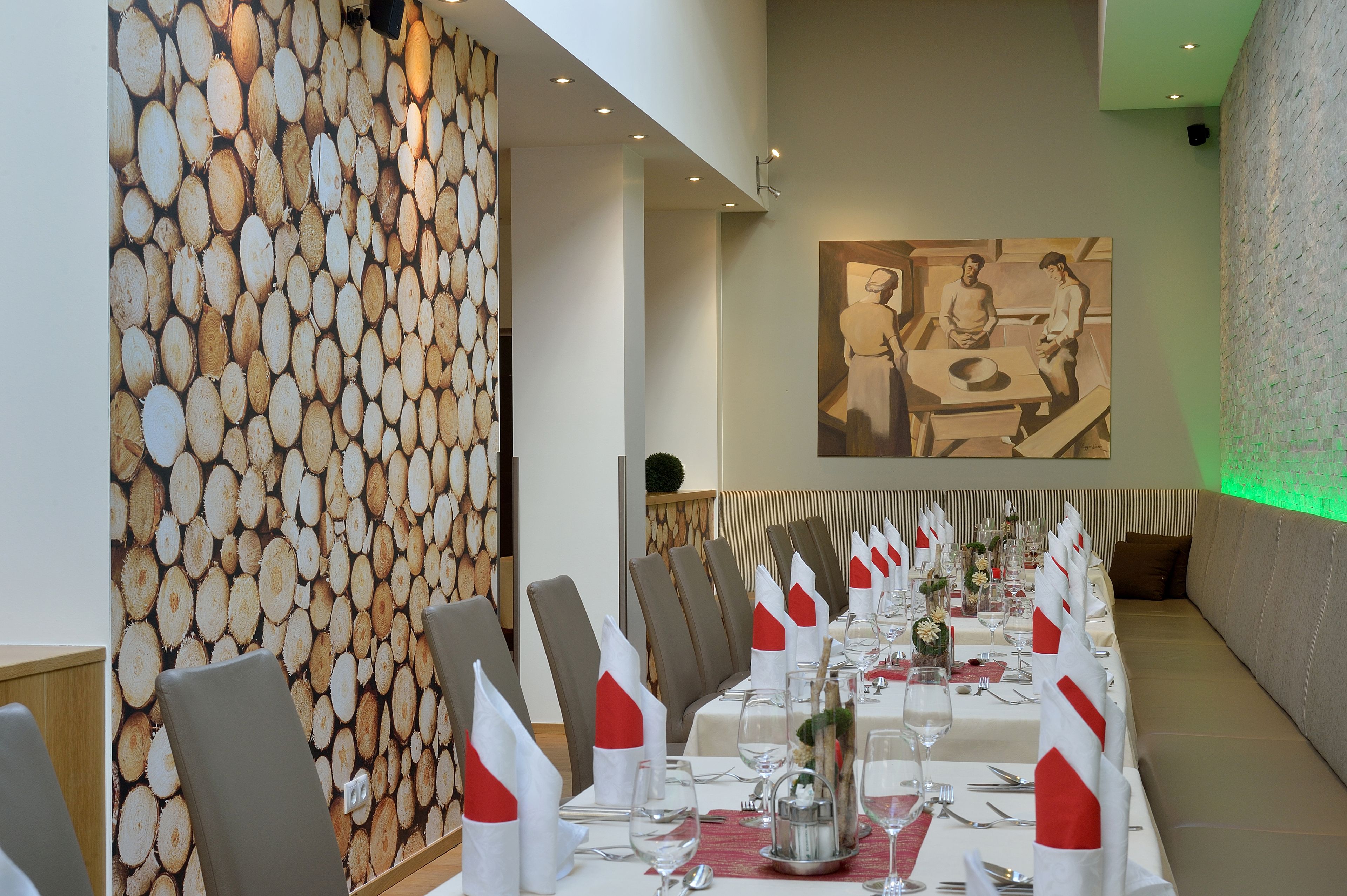 A long dining table set with white napkins, silverware, and glassware, flanked by chairs and a wall with a wood log pattern.