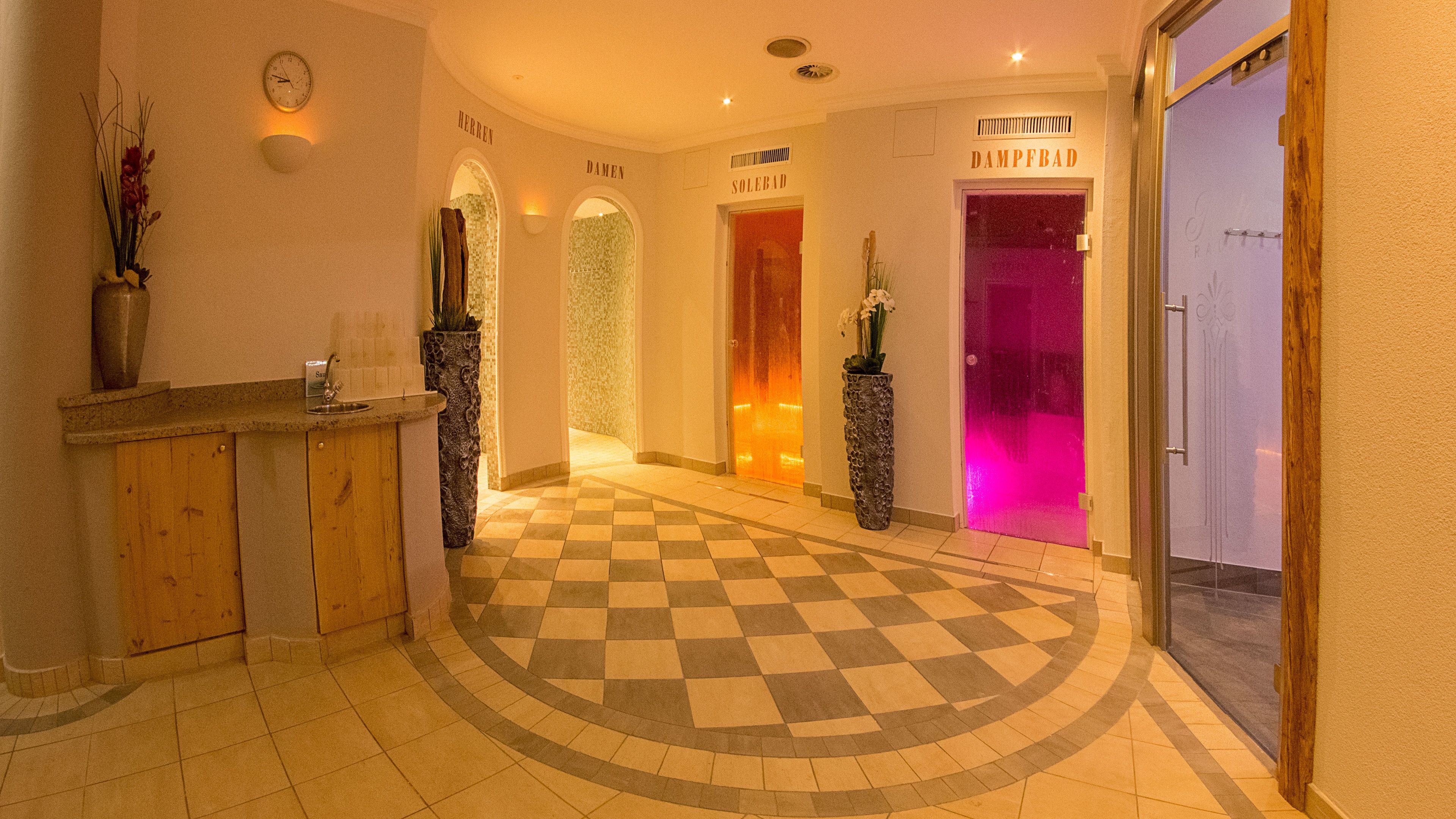 Un vestíbulo de hotel con suelo a cuadros, un mostrador de recepción y puertas con los letreros "Damen" y "Dampfbad".