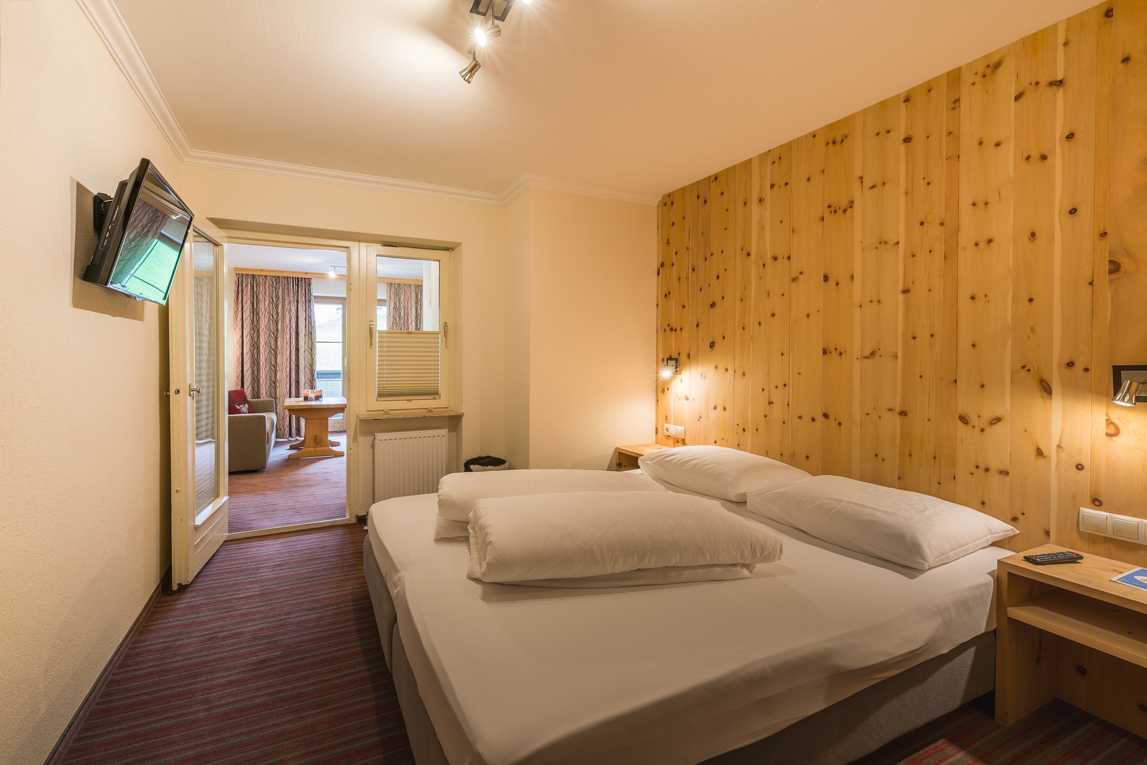 Habitación de hotel con cama, televisor montado en la pared, cabecero de madera y una mesita de noche pequeña.