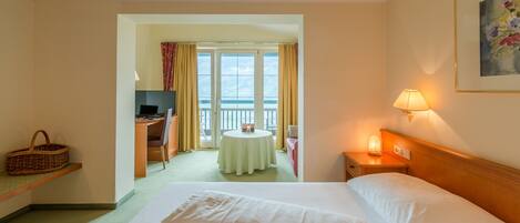 Quarto de hotel com cama, escrivaninha, cadeira, mesa, televisão, abajur e janela com cortinas.