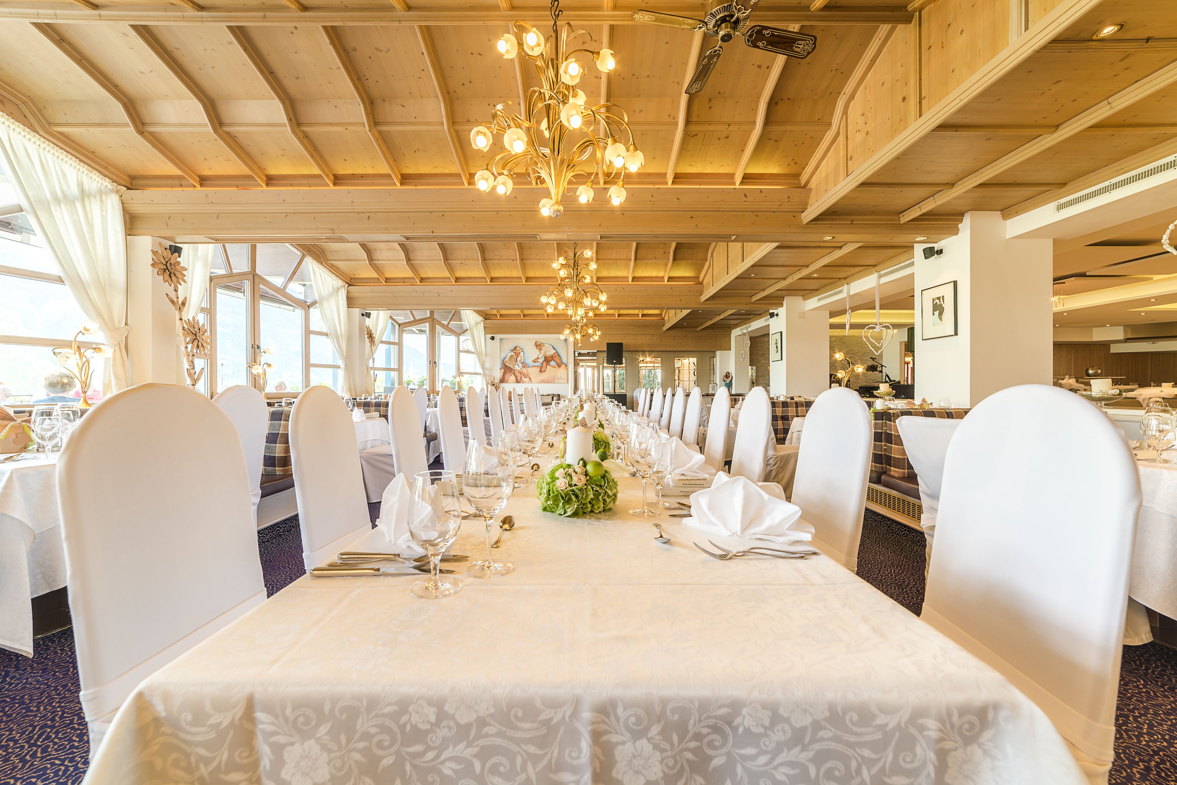 Une salle de banquet avec de longues tables dressées pour un événement formel, agrémentée d’un lustre et de poutres en bois apparentes au plafond.