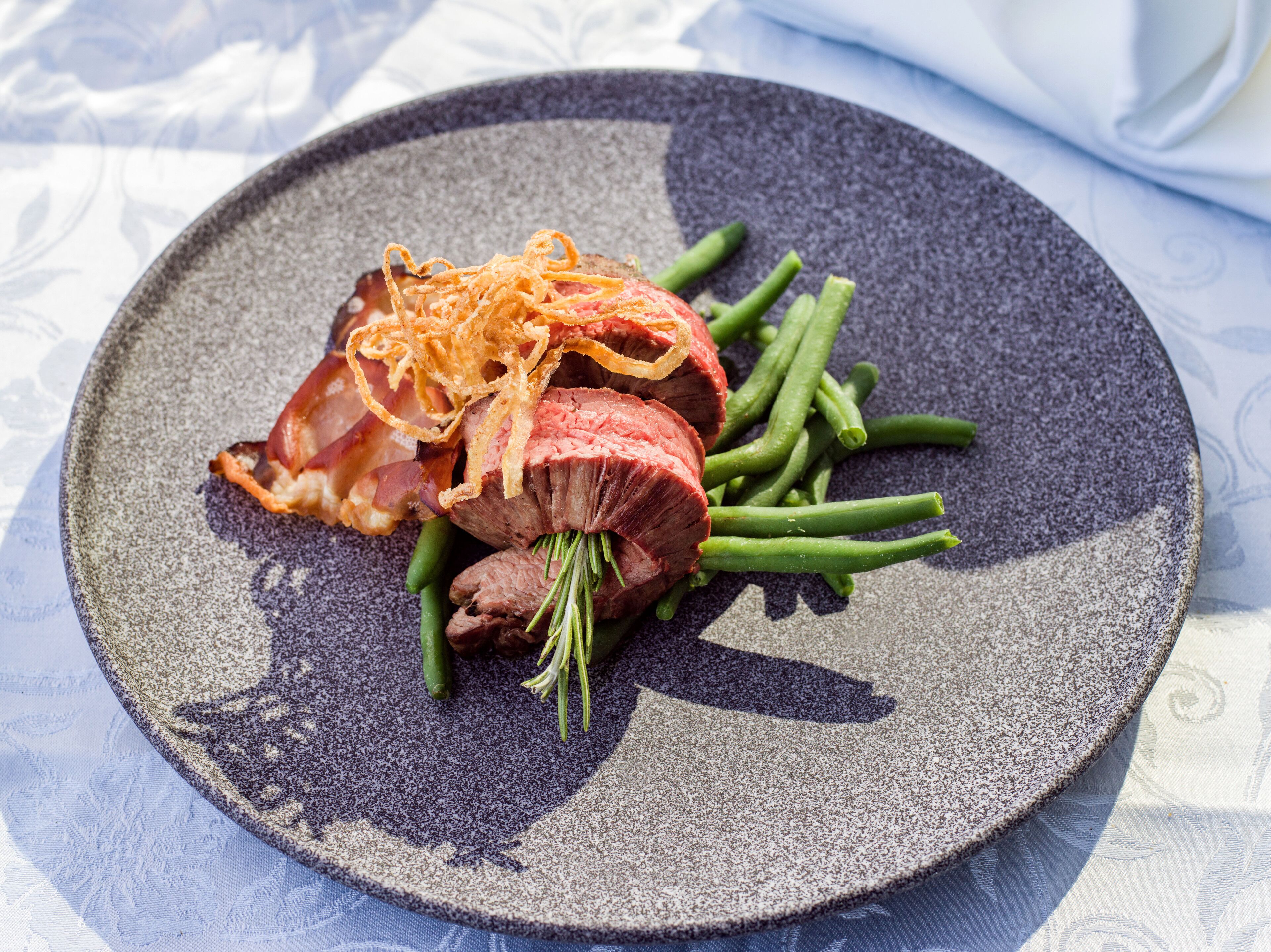 A plate with sliced meat, green beans, and crispy garnish on a textured plate.