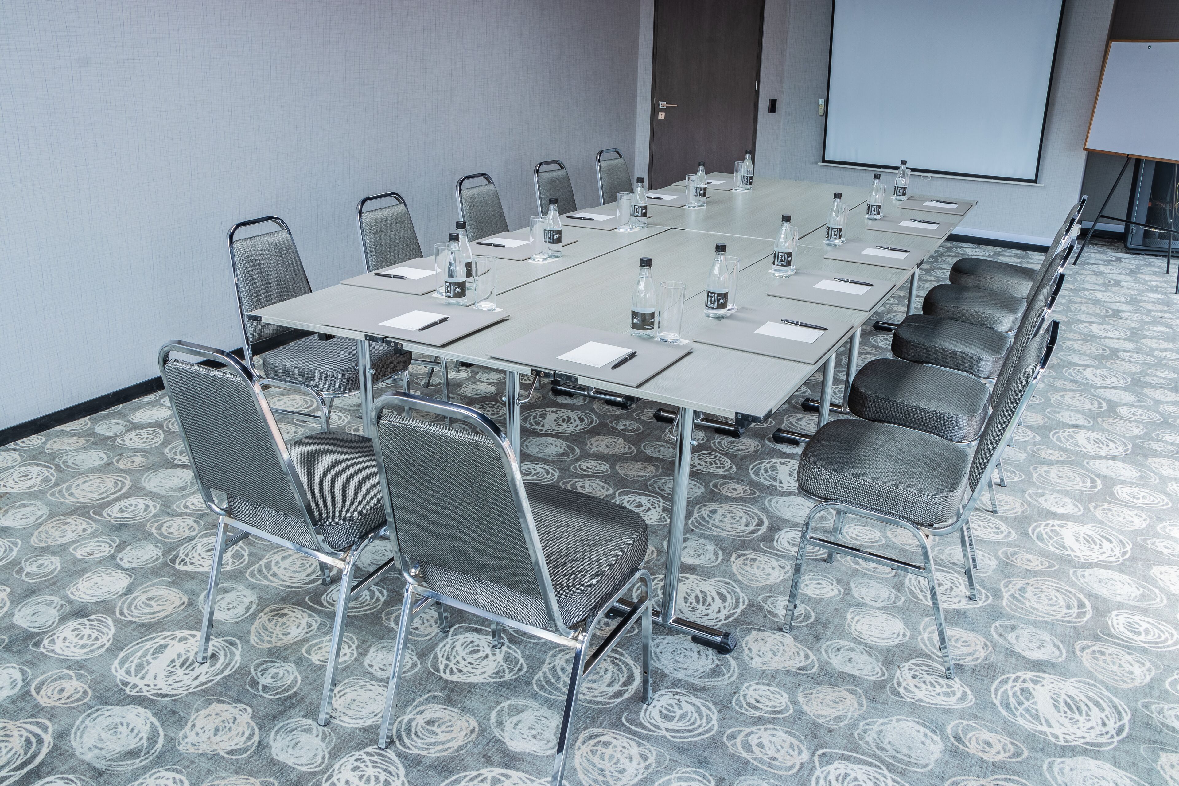 A conference room with a long table, chairs, water bottles, and a projector screen.