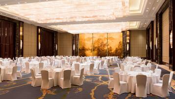 Un amplio salón de banquetes con mesas redondas y sillas blancas, una alfombra estampada y una gran lámpara de araña.