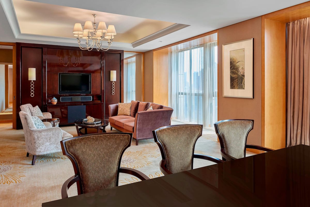 A hotel room with a large flat-screen TV, a sofa, armchairs, a dining table, and a chandelier.