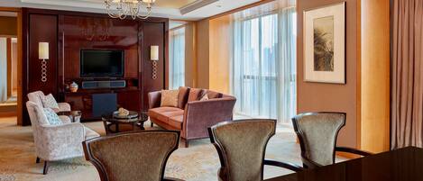 A hotel room with a large flat-screen TV, a sofa, armchairs, a dining table, and a chandelier.