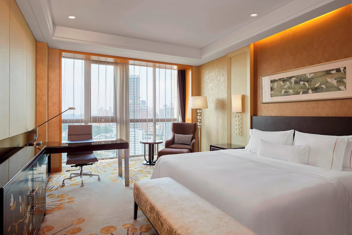 A hotel room with a large bed, a desk with a chair, a small table, and a chair. There is a window with a view of buildings.