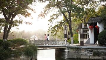 A traditional bridge over a calm river, surrounded by lush greenery and a small traditional building with red lanterns.