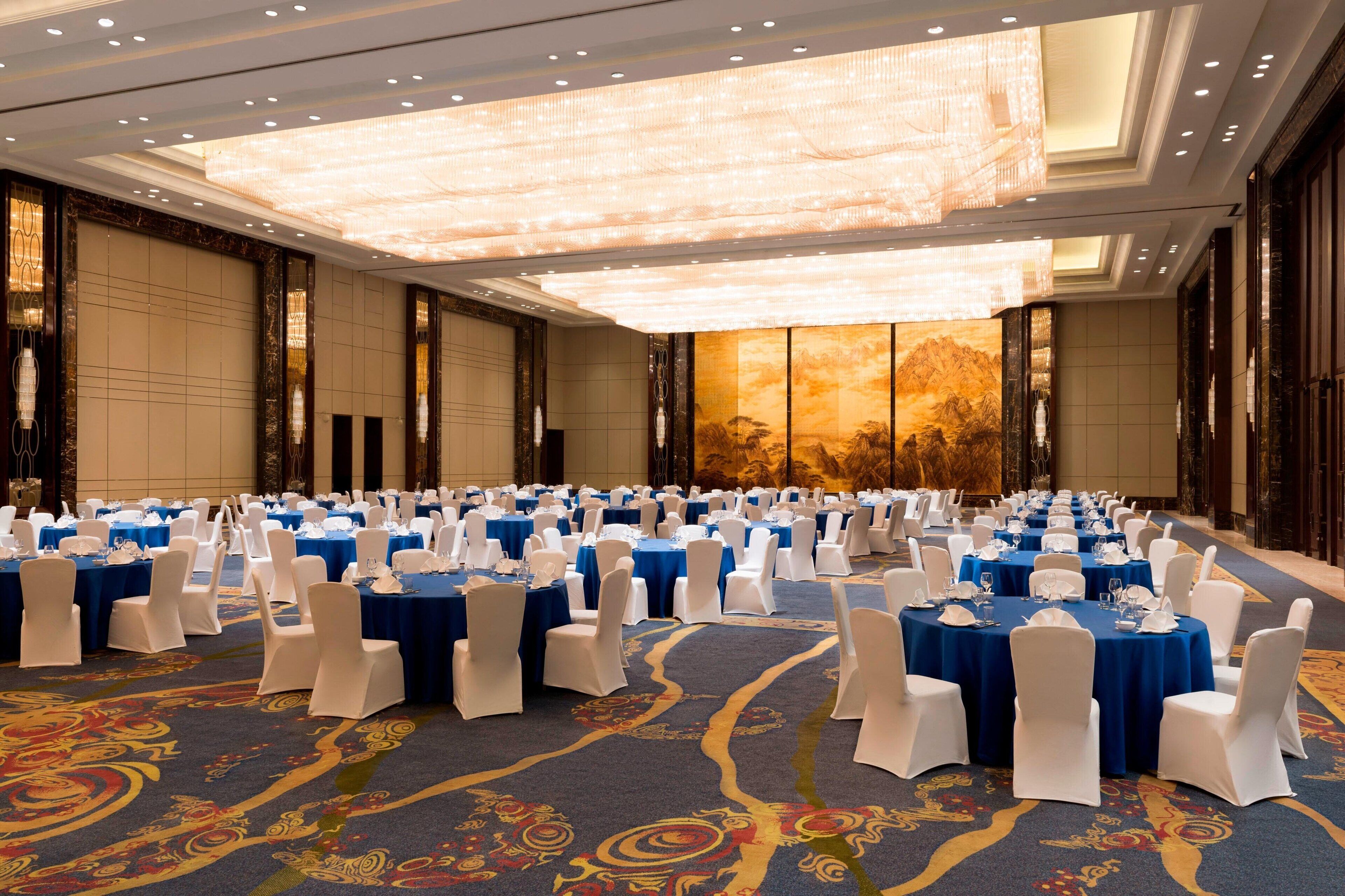 A banquet hall with round tables, white chairs, and a large chandelier.