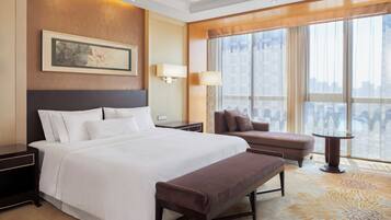A hotel room with a large bed, a bench, a nightstand, a lamp, a wall-mounted artwork, and a large window with a view.