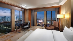 A hotel room with a large bed, a desk, a chair, and a view of a cityscape at night.