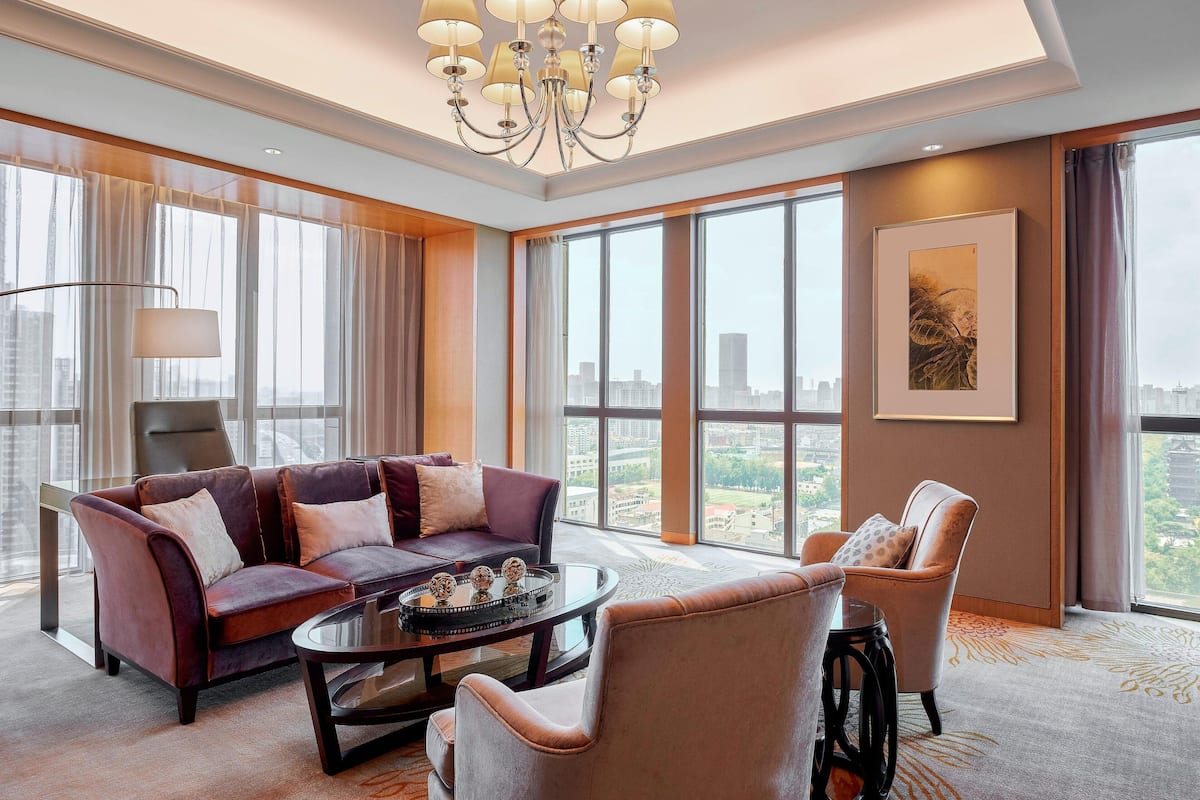 A modern hotel room with a large window offering a city view, a plush sofa, a round coffee table, and a chandelier.