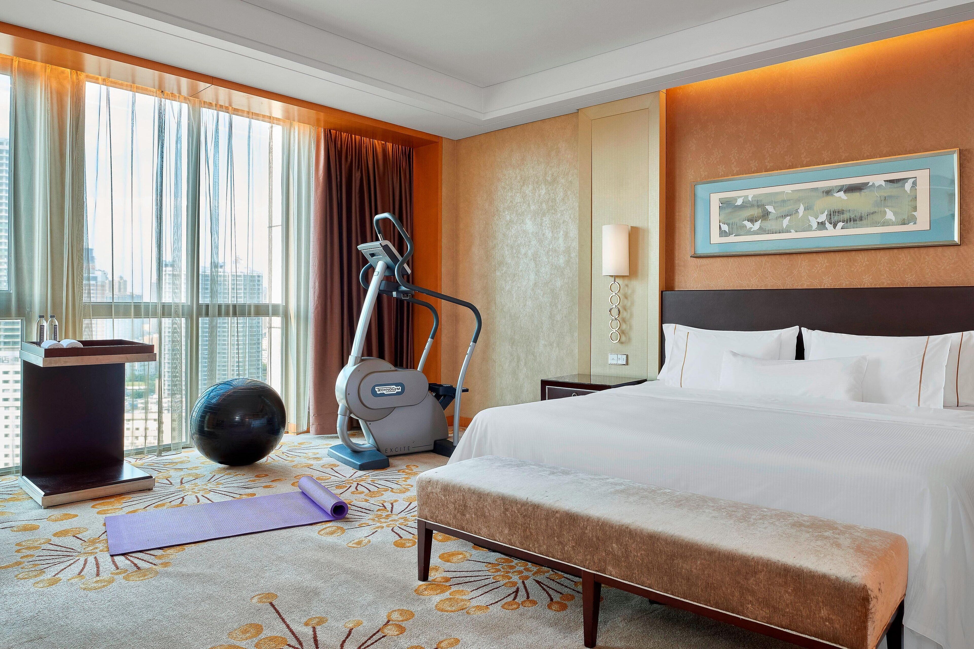Habitación de hotel con una cama grande, equipo de ejercicio, esterilla de yoga y vistas a la ciudad a través de amplios ventanales.