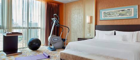 Habitación de hotel con una cama grande, equipo de ejercicio, esterilla de yoga y vistas a la ciudad a través de amplios ventanales.