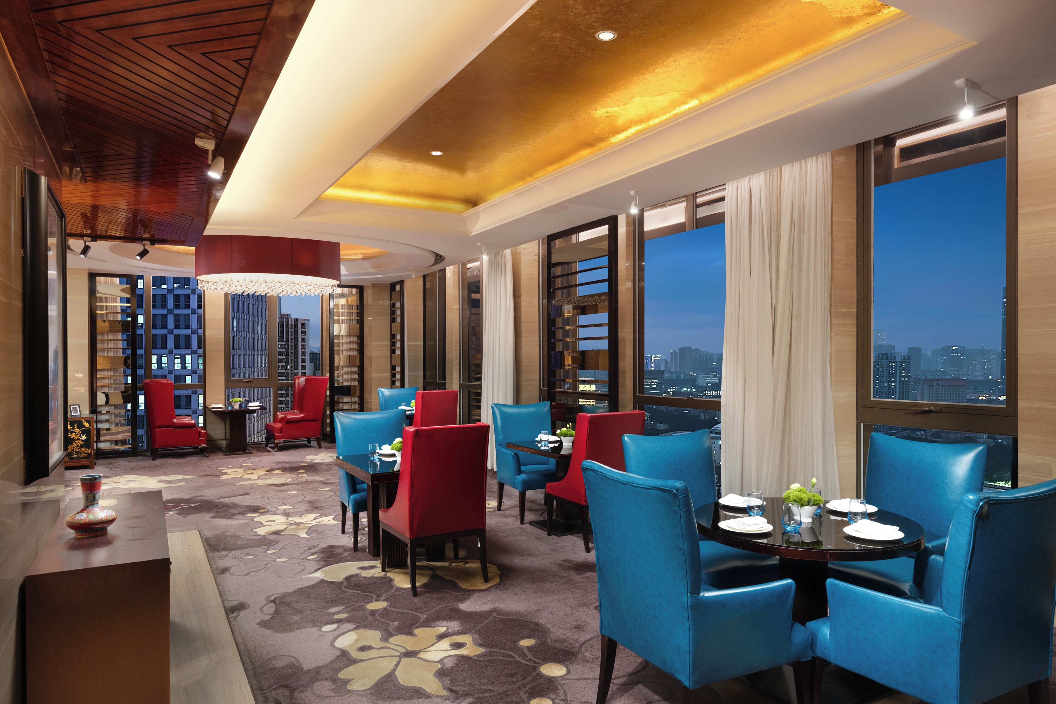 Un vestíbulo de hotel con diseño moderno, que cuenta con un amplio comedor con sillas azules y rojas, una araña y grandes ventanales con vistas a la ciudad.