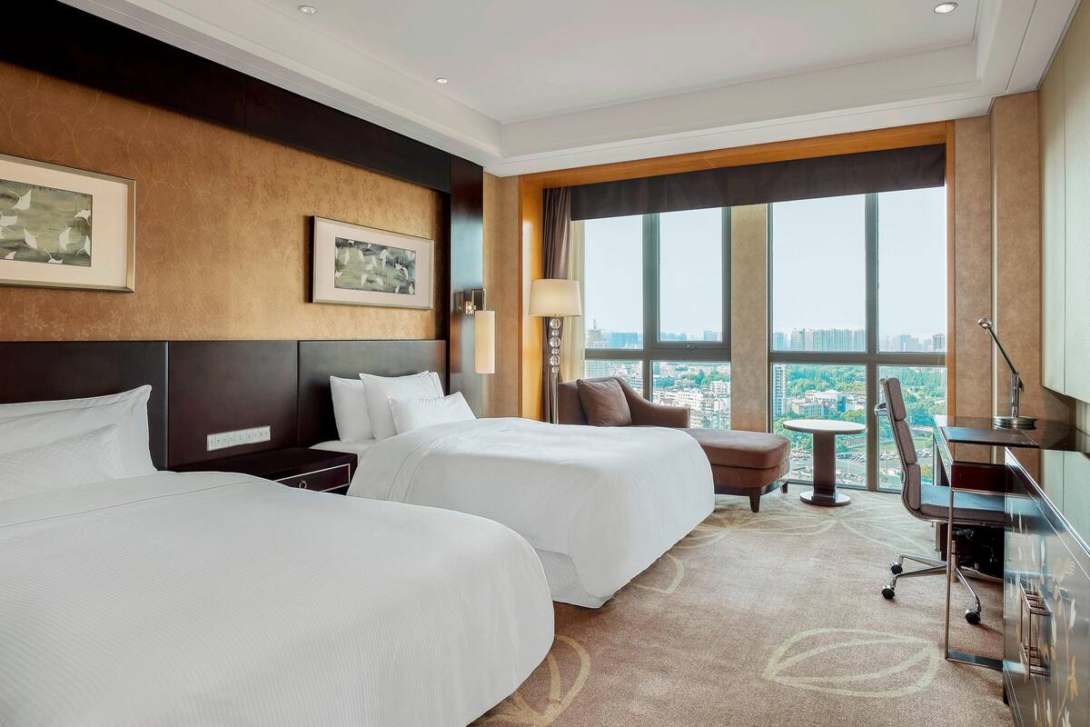 A hotel room with two beds, a desk, a chair, a lamp, and a view of the city.