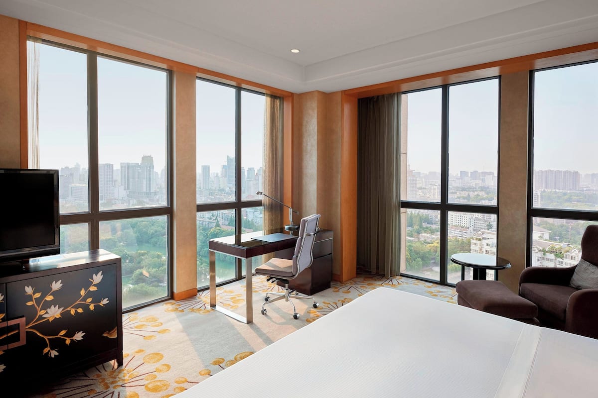 A hotel room with a large bed, a desk with a chair, a TV, and a view of the city through large windows.