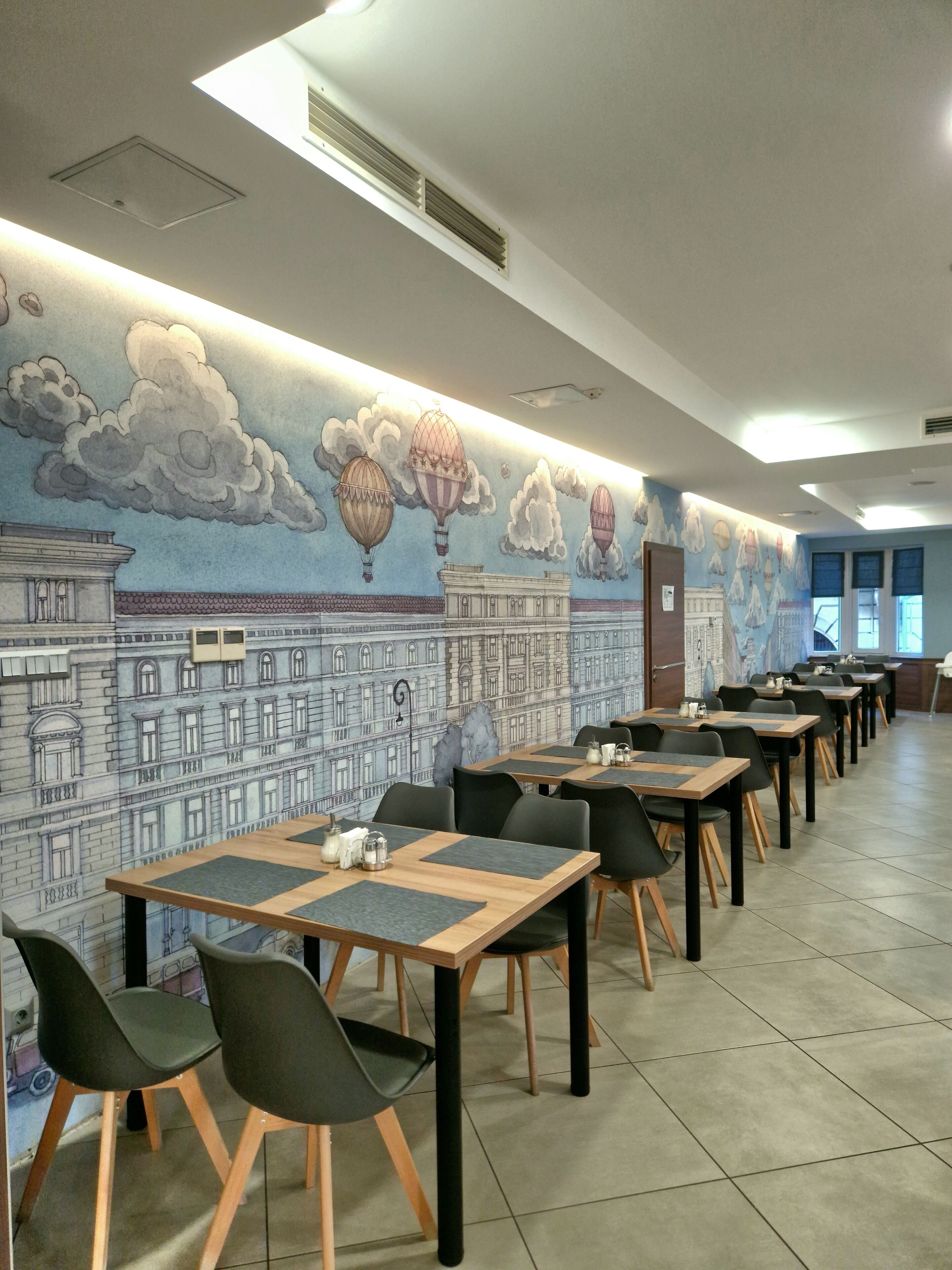Un espace repas avec des tables et des chaises, une fresque représentant des bâtiments et des montgolfières sur le mur, et une porte au fond.
