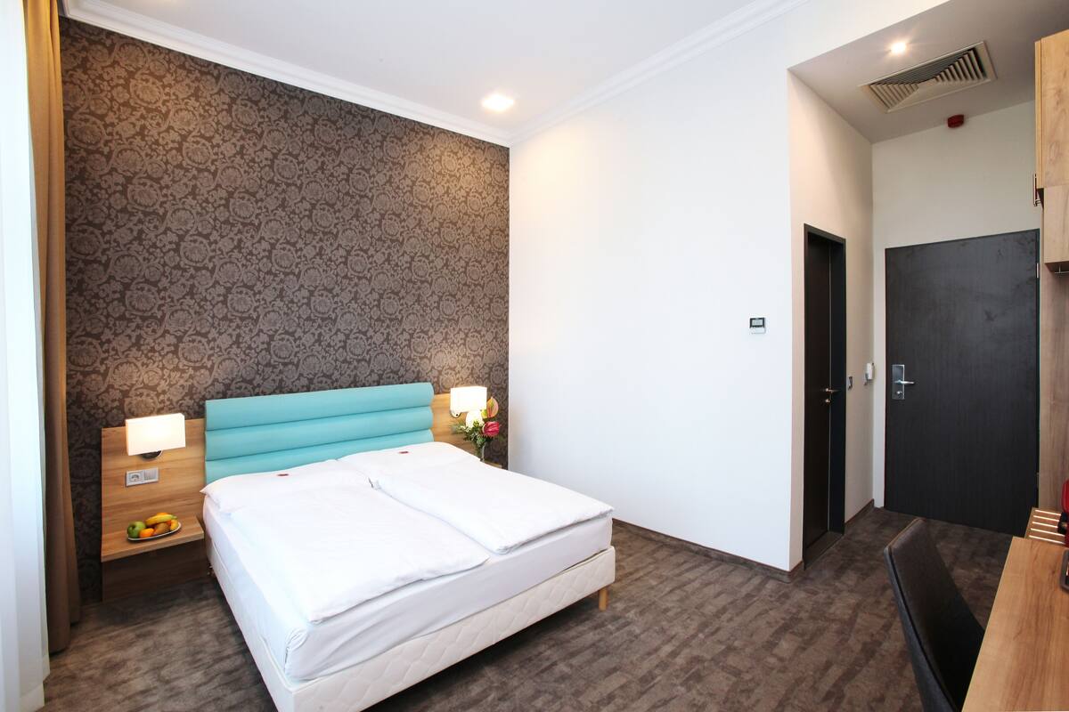 Une chambre d’hôtel avec un lit, une table de chevet avec une lampe, une tête de lit en bois, un papier peint à motifs, une porte sombre et une petite table avec une chaise.