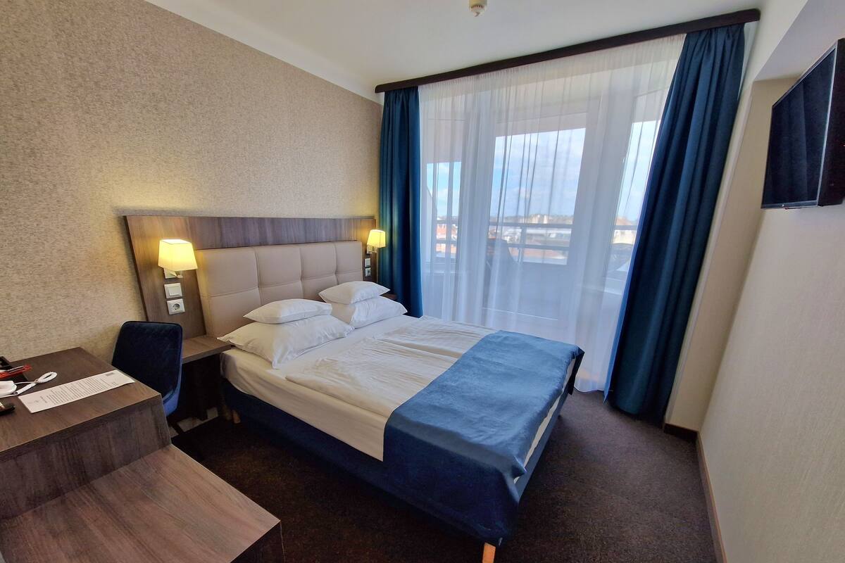 Une chambre d’hôtel avec un lit, un bureau et une chaise. Il y a une lampe sur le bureau et une fenêtre avec des rideaux.