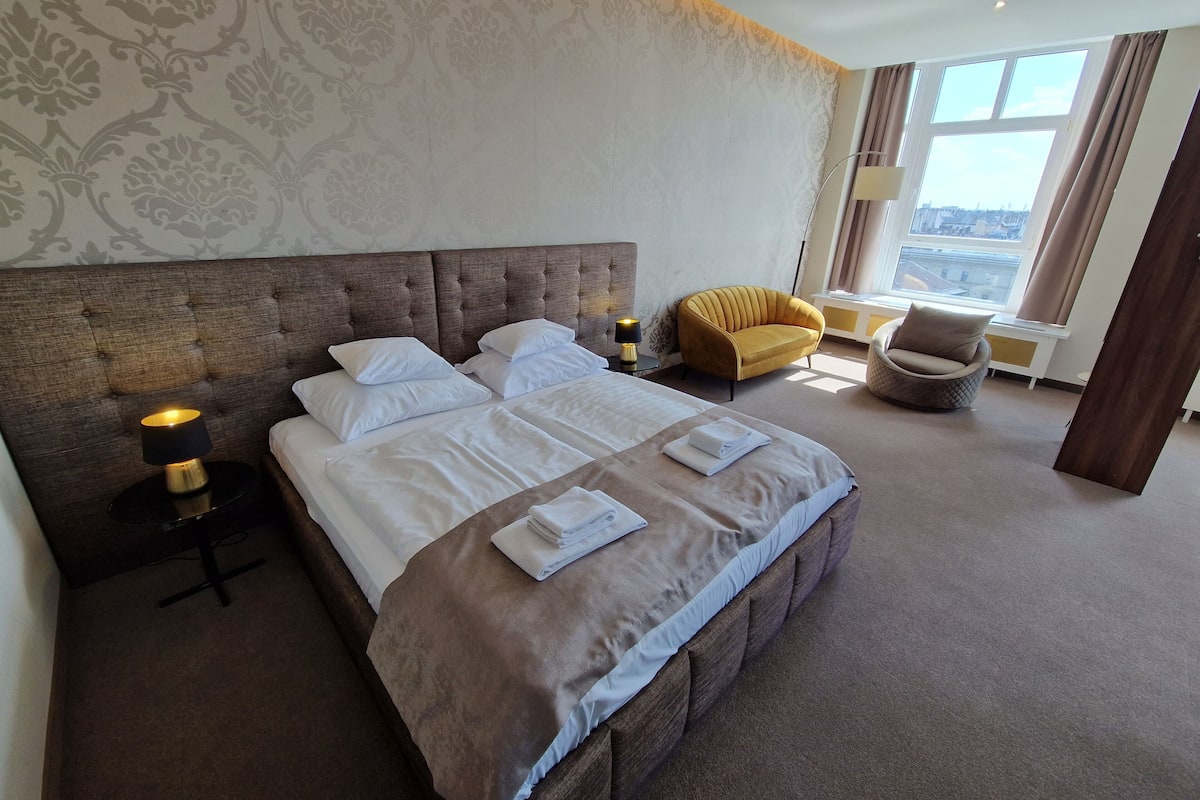 Une chambre d’hôtel avec un grand lit, une chaise, une lampe et une vue sur la ville par la fenêtre.