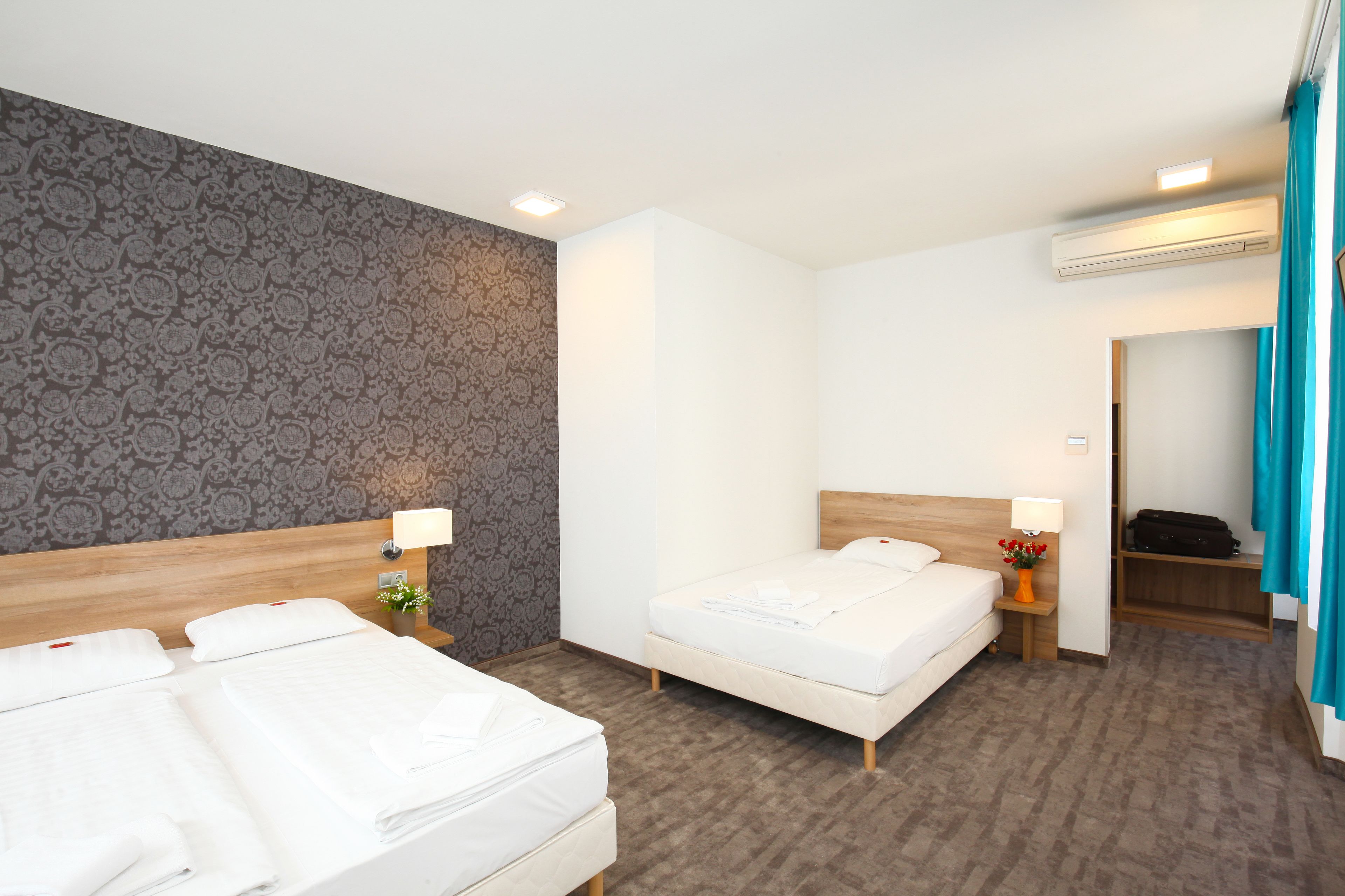 A hotel room with two beds, a wooden headboard, a patterned wall, a small table with a lamp, and an air conditioning unit.