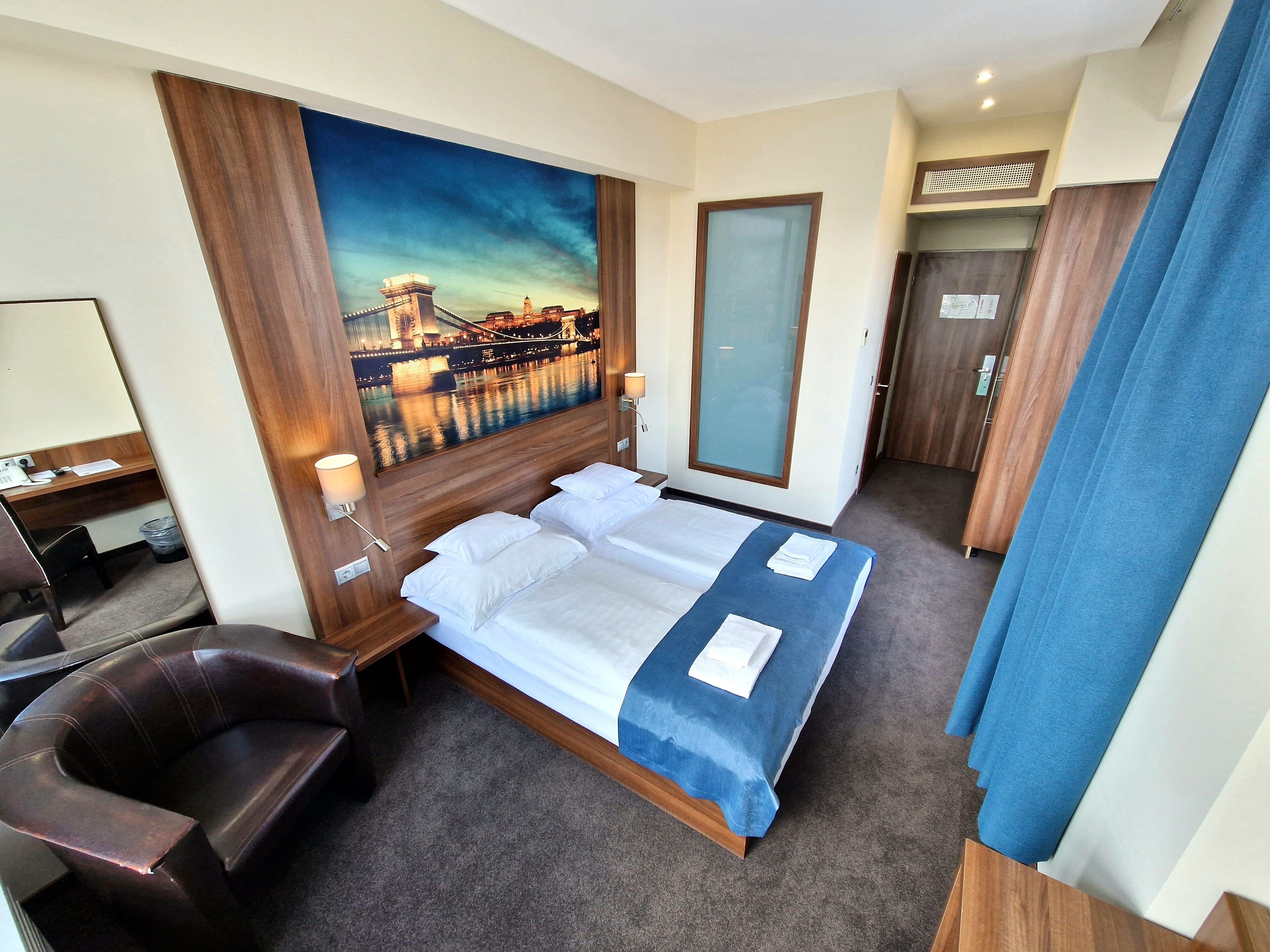 A hotel room with a bed, a desk, a chair, a large window, and a painting of a cityscape.