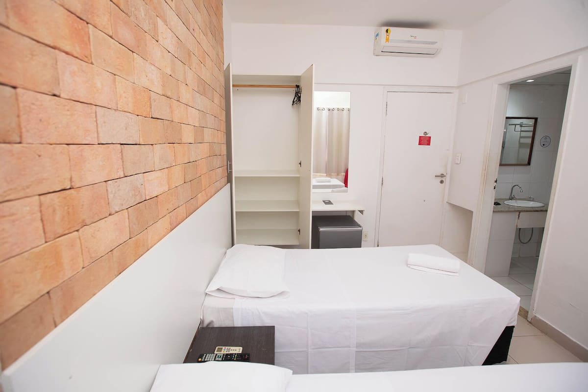 Ein Hotelzimmer mit Backsteinwand, einem Bett mit weißen Laken, einem kleinen Tisch mit Fernbedienung und einem Badezimmer mit Waschbecken und Spiegel.