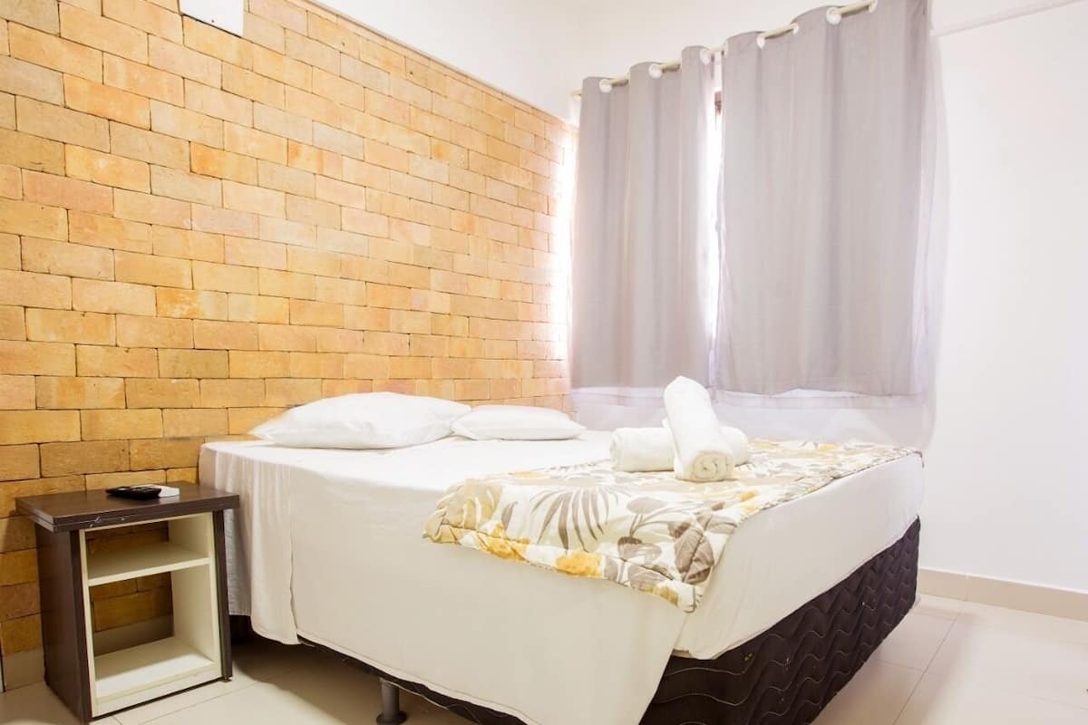 Ein Hotelzimmer mit Backsteinwand, einem Bett mit weißer Bettdecke und Kissen, einem Nachttisch mit Fernbedienung und einem Fenster mit Vorhängen.