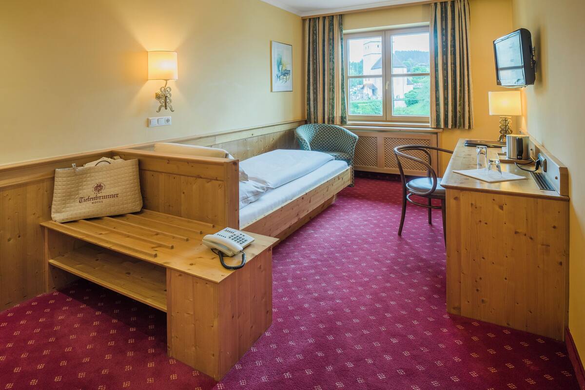 A compact hotel room with a wooden bed, a small desk, a chair, a window with curtains, and a television.