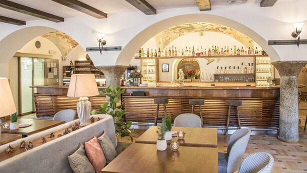Una zona de bar con mostrador de madera y estantes llenos de botellas, rodeada de asientos y un pilar de piedra.