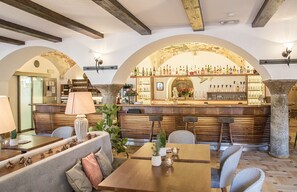 Uma área de bar com balcão de madeira e prateleiras cheias de garrafas, rodeada por assentos e um pilar de pedra.