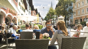Un café al aire libre concurrido, con sillas y mesas de mimbre, rodeado de edificios históricos con señalización visible.
