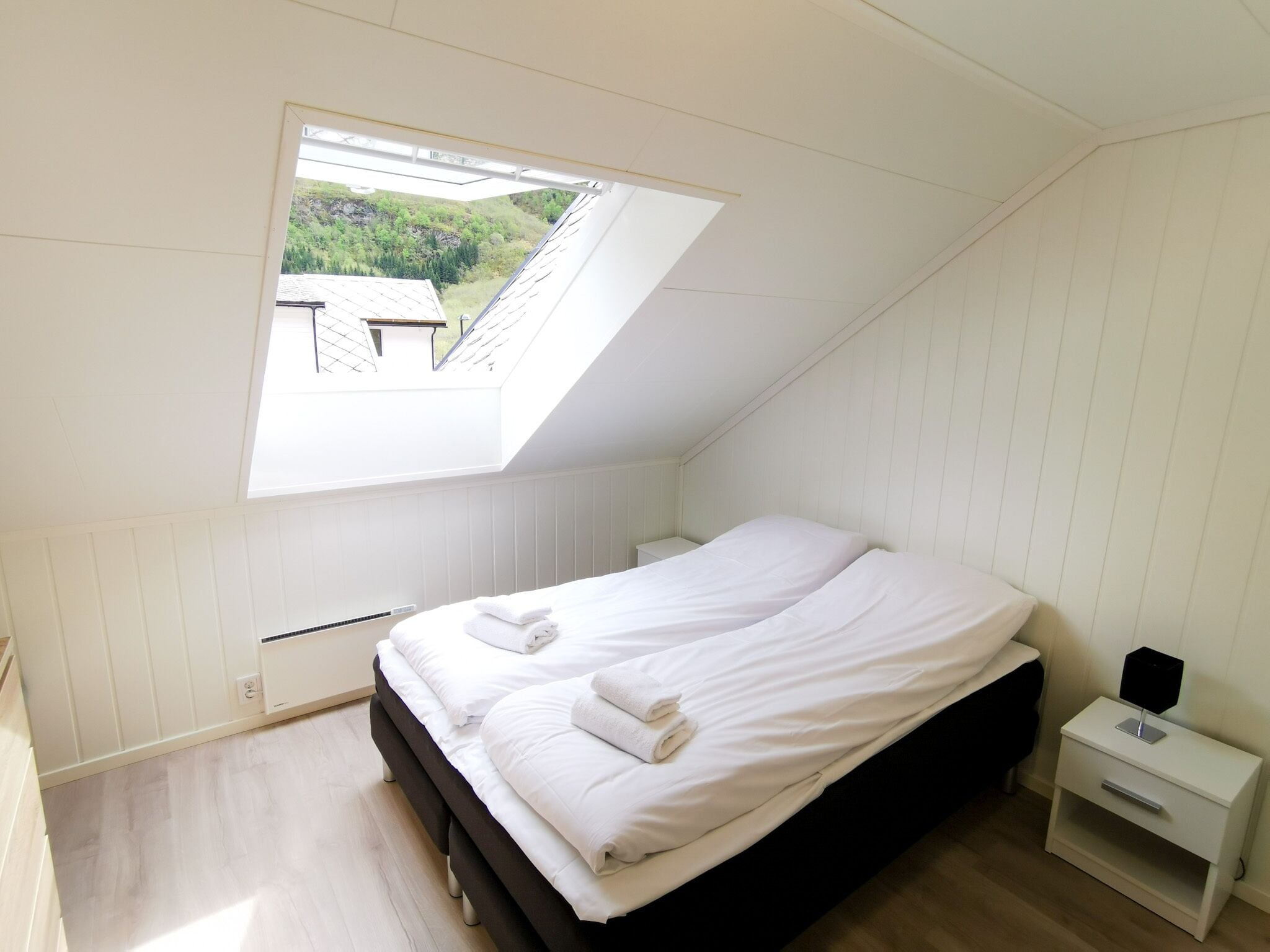 Ein Schlafzimmer mit schrägen Wänden, einem Oberlicht, einem Bett mit weißen Leinen, einem Nachttisch mit Lampe und Blick auf Grünflächen.