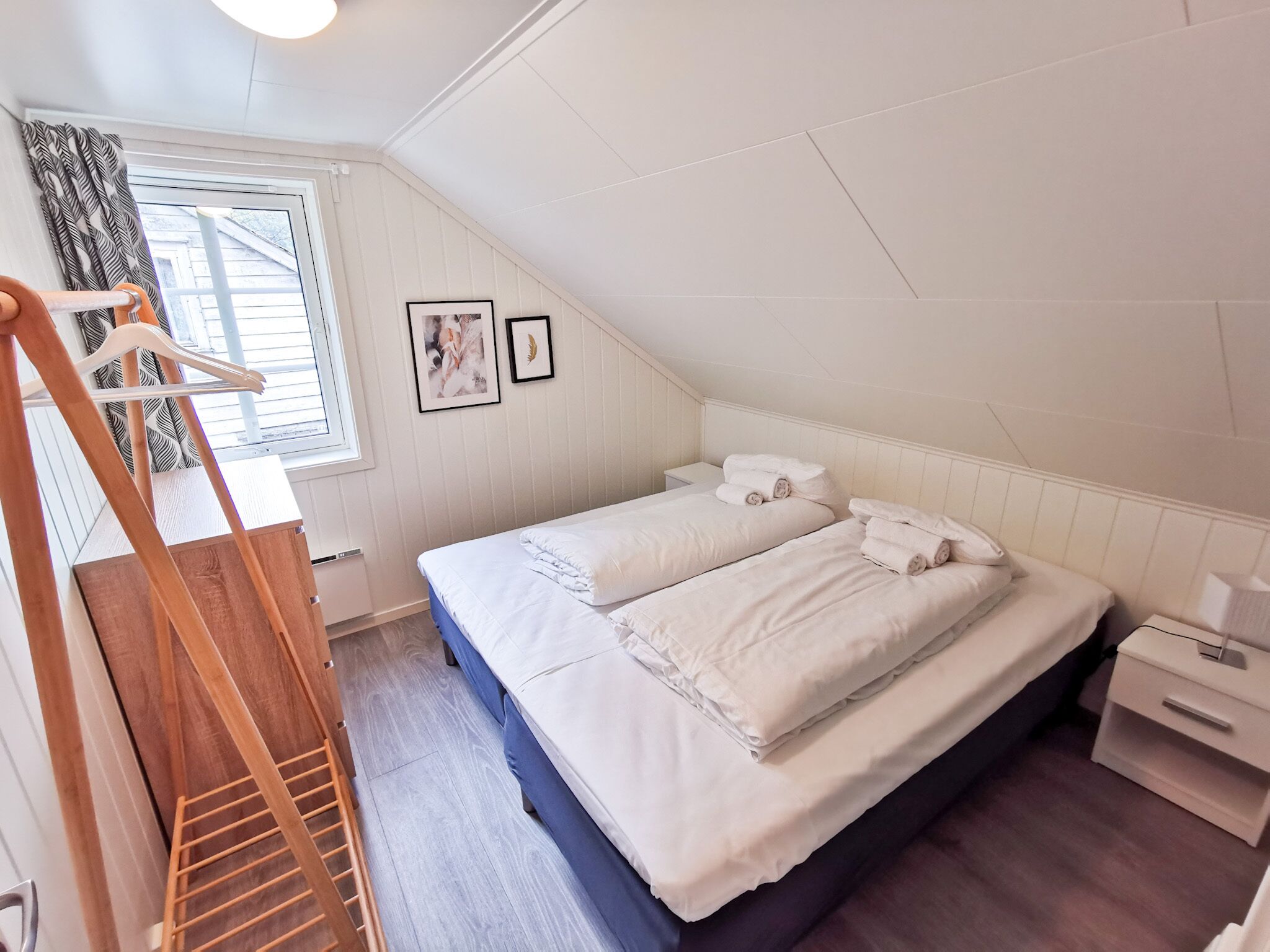Een slaapkamer met een bed, een nachtkastje, een houten ladder, een raam met gordijnen en twee ingelijste foto's aan de muur.